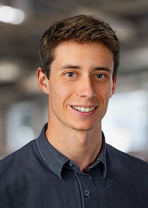 Portrait picuture of Maximilian Mali. Smiling with blurred background wearing a blue shirt in casual style.